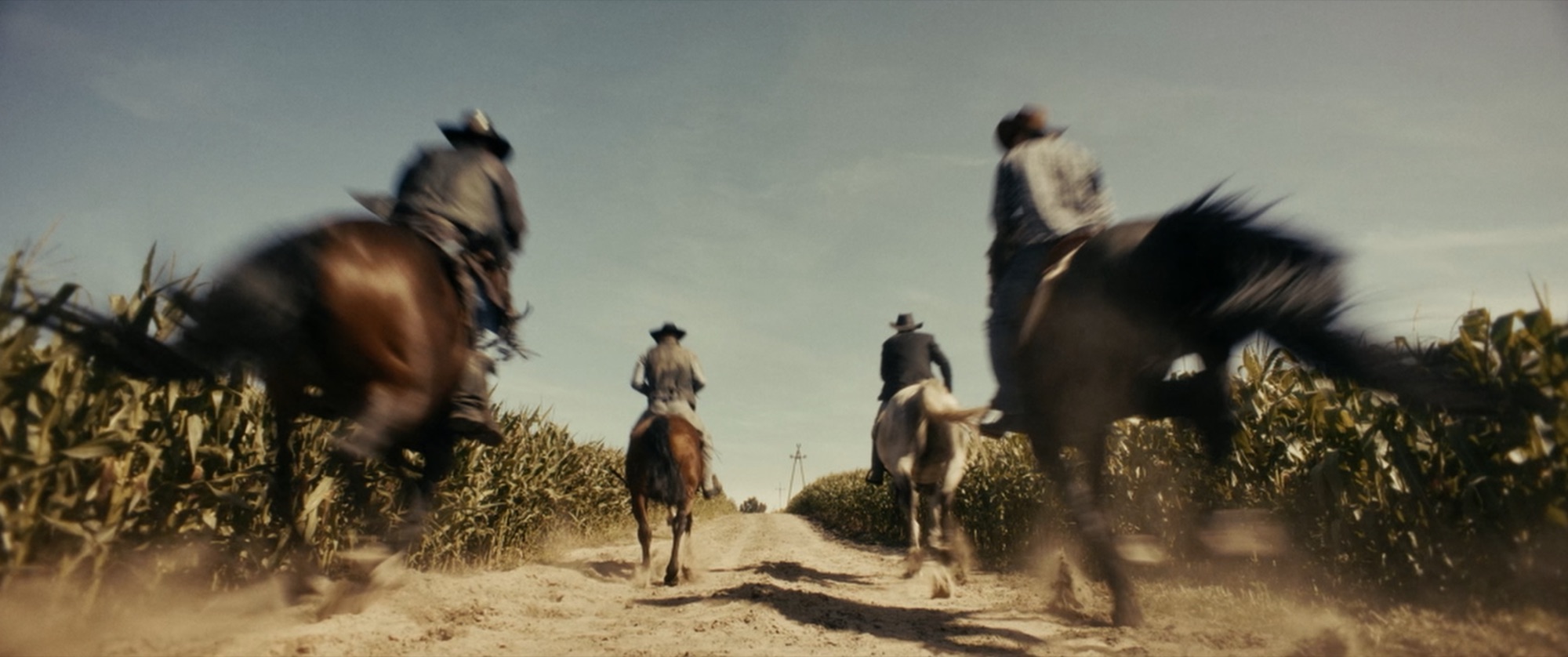 Low rear view of two riders galloping down a dusty track between tall cornfields, heavy motion blur - Amon Amarth